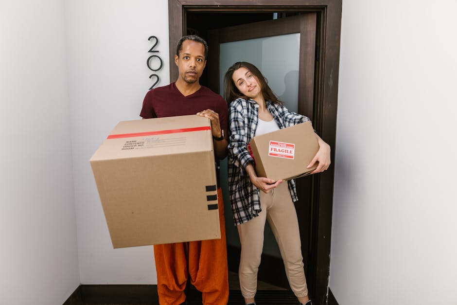 A young woman sitting on a wooden floor inside a property, surrounded by numerous large cardboard boxes of various sizes, some sealed with red tape and others open. She is wearing casual clothing, inc