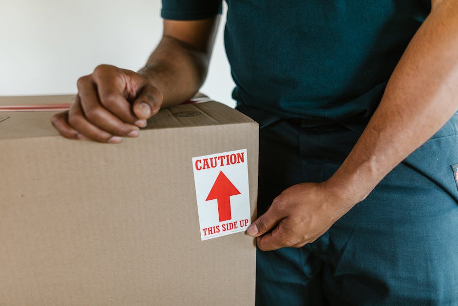 A person wearing a dark blue T-shirt and blue trousers is seen during the packing process inside a property, holding a large cardboard box with both hands. The box has a red and white caution label th