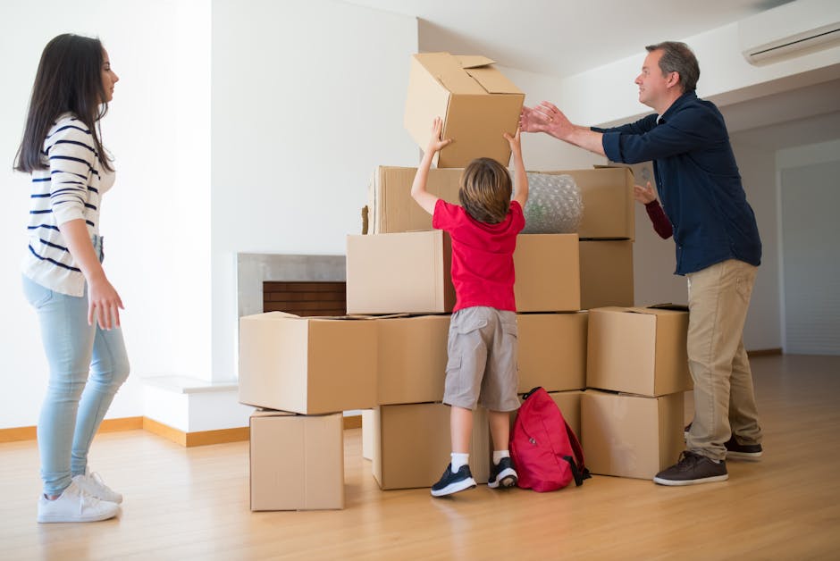 A person wearing a dark blue T-shirt and blue trousers is seen during the packing process inside a property, holding a large cardboard box with both hands. The box has a red and white caution label th
